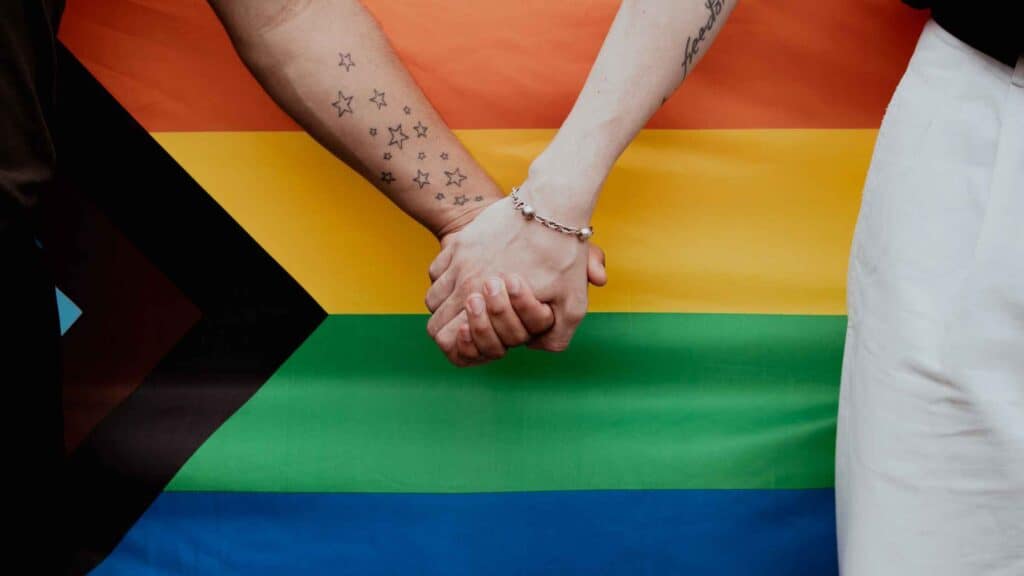 Two people holding hands with a rainbow flag in the background, symbolizing gender affirmation and s.